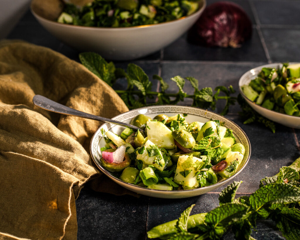 A close-up of a fresh green salad in a decorative bowl, garnished with mint leaves and mixed vegetables, set against a rustic table with a cloth and additional salad in the background.